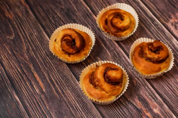 Dört taze pişmiş tarçınlı rulo beyaz kağıt bardaklarda koyu ahşap bir yüzeye dizilmiş..