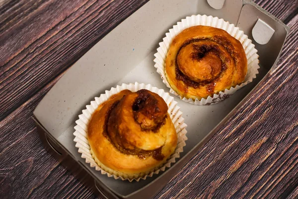 Kağıt bardakların içinde kıvrımlı iki tatlı tarçın rulosu, koyu ahşap bir yüzeyde karton bir kabın içine konur..