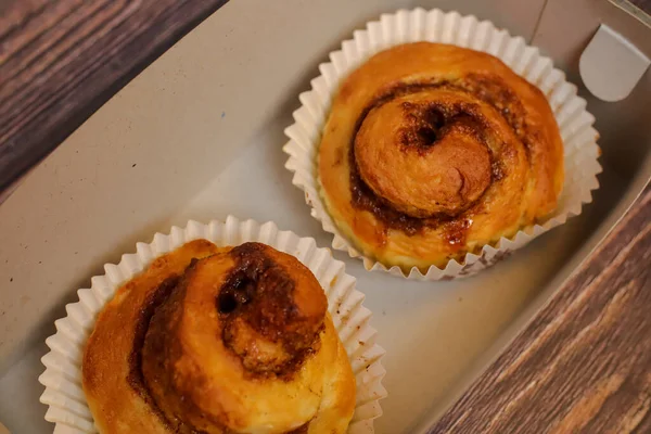 İki yeni pişmiş tarçın rulosu açık gri bir karton kutunun içinde beyaz kağıt bardaklarda sunuluyor..