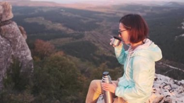 Gezgin yürüyüşçü genç kadın termostan çay içiyor ve yaz vadisinin arka planındaki dağın tepesinde dinleniyor..