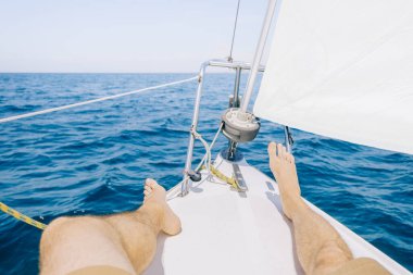 Tanımlanamayan genç adam, güneşli bir yaz gününde mavi denizde yelken açan yatın pruvasında dinleniyor, bacakları görüyor. Bakış açısı.