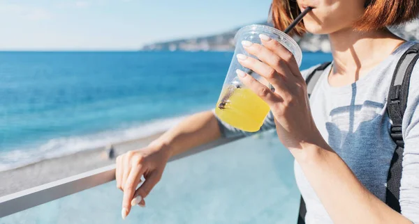 美丽的年轻女子在夏天在大海的背景下微笑着喝着新鲜的果汁 把注意力放在手边 — 图库照片