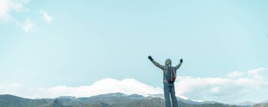 Elleri havada gezgin, sırtını kameraya dayamış bir dağ zirvesinin arka planında duruyor..