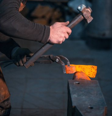 Demirci maşayla dövülen kızgın bir metal parçasını elinde tutuyor..