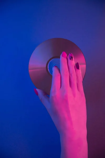 Γυναικείο χέρι κρατά ένα συμπαγές δίσκο σε μπλε φόντο σε μωβ νέον φως. — Φωτογραφία Αρχείου