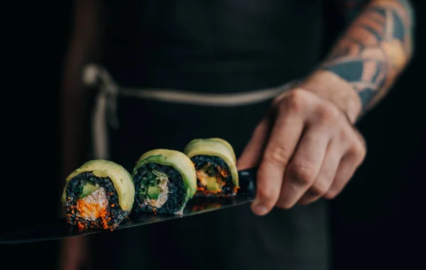 Hombre chef sostiene rollos de sushi en un cuchillo. — Foto de Stock