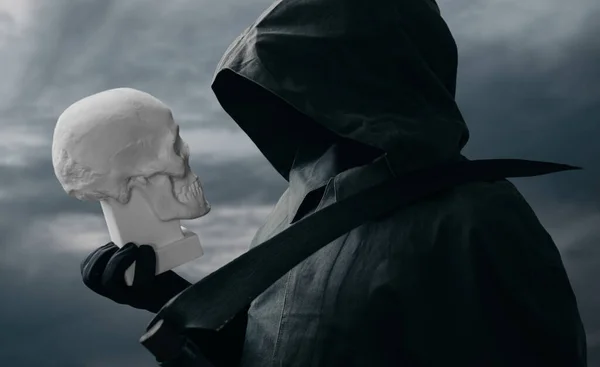 The grim reaper stands with a scythe in the background of the dark sky and looks at the skull. — Stock Photo, Image