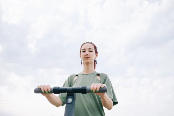 Young millennial woman stands on a scooter and looks at the camera. — Stock Photo, Image