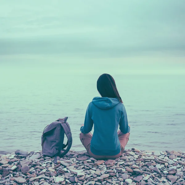 Meisje met rugzak zittend op stenen strand — Stockfoto