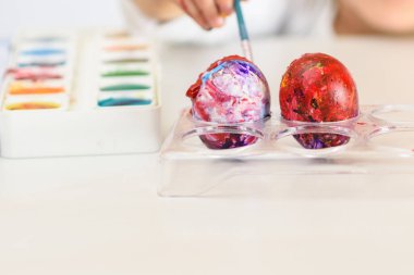 Child hands painted Easter eggs, paints and brushes on white table. Preparation for the Easter. Kid hands draw with colors.