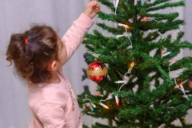 Sevimli küçük bebek evde kırmızı cam süslemelerle bir Noel ağacını süslüyor..