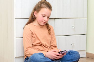 Kız yerde oturuyor ve akıllı telefondaki Webinar 'ı izliyor şeftali rengi bir tişört giyiyor..