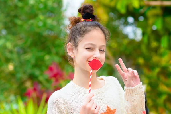 Verrast knipogen klein meisje houden grappige lippen op stok en tonen overwinning gebaar op groene natuur achtergrond. — Stockfoto