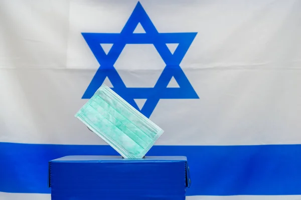 Вибори в Ізраїль. Маска медичного обличчя в ящику для голосування на тлі прапора Ізраїлю.. — стокове фото