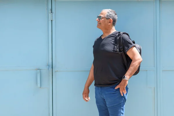 Усміхнений старший чоловік стоїть перед синіми дверима в місті, тримаючи його чорний рюкзак, дивлячись в бік . — стокове фото