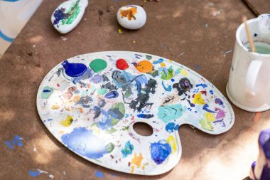 Çocuklar taşların üzerine boya sürerek yaratıcı faaliyetlerde bulunurlar. Renkli bir palet, boyalı taşlar ve sanat malzemeleriyle çevrili bir masada yatıyor..