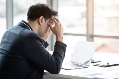 Stresli iş adamı dizüstü bilgisayarla çalışıyor ve ofis odasındaki kayıplardan sonra baş ağrısı çekiyor..