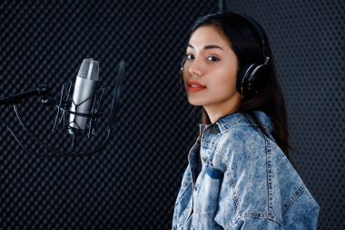 Profesyonel bir stüdyoda mikrofon önünde şarkı kaydeden, kulaklık takan genç Asyalı kadın vokalistin mutlu bir gülümsemesi.