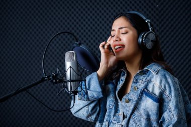 Profesyonel bir stüdyoda mikrofon önünde şarkı kaydeden, kulaklık takan genç Asyalı kadın vokalistin mutlu bir gülümsemesi.