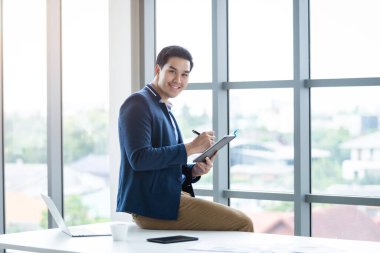 Asyalı genç iş adamının iş planı defterine ve dizüstü bilgisayarına kaydedilen notu okuduğunu düşünen akıllı telefon masanın üstünde, ofis odasının arka planında oturuyor..