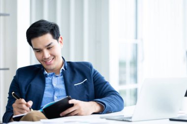 Mutlu bir ruh hali içinde Asyalı genç bir iş adamı ofis odasındaki dizüstü bilgisayara ve not defterine notlar yazarak çalışıyor..