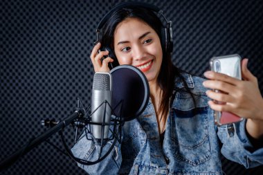 Genç Asyalı kadın vokalistin mutlu bir gülümsemesi kulaklık takıyor, akıllı telefonuyla selfie çekiyor profesyonel bir stüdyoda mikrofon önünde şarkı kaydediyor.