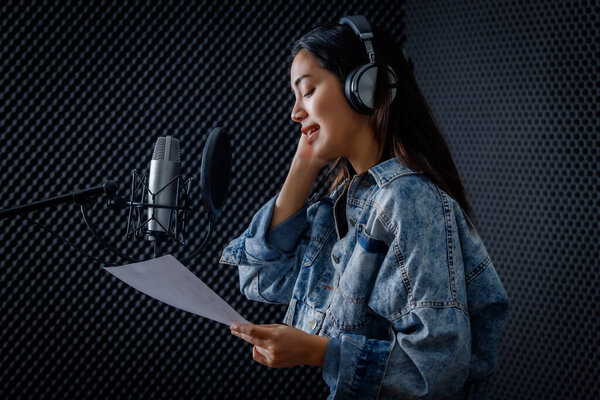 Happy cheerful pretty smiling of portrait a young Asian woman vocalist Wearing Headphones recording a song front of microphone in a professional studio