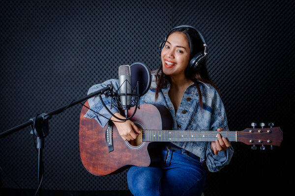 Happy cheerful pretty smiling of portrait a young Asian woman vocalist Wearing Headphones with a Guitar recording a song front of microphone in a professional studio