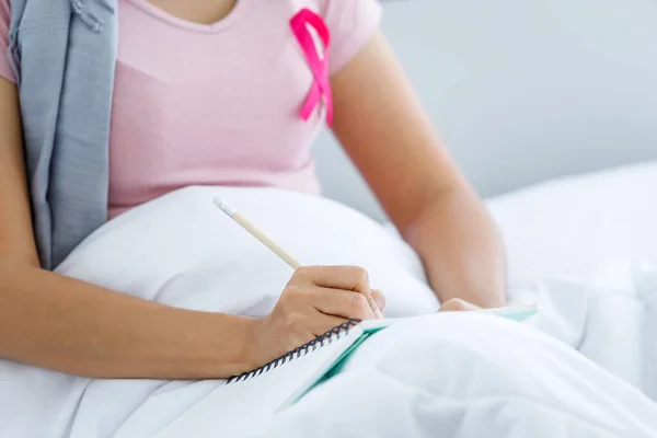 一位亚洲女性患乳腺癌的患者 带着粉色丝带 头戴头巾接受化疗后 在床上的笔记本里记笔记在家里的卧室里 医疗保健 药物上 — 图库照片