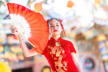 Güzel bir portre gülümsüyor. Küçük şirin bir Asyalı kız. Geleneksel Çin cheongsam süslemesi takıyor ve Çin Yeni Yıl Festivali için Çin tapınağında Fanning düzenliyor.