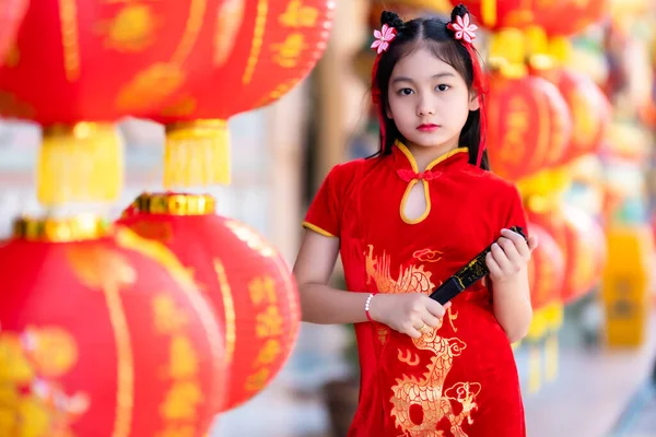 赤い伝統的な中国のチョンサムを身に着けている小さなアジアの女の子とそれに書かれた中国語のアルファベットの祝福とFanningandランタンを保持新年のための幸運祝福の賛辞の装飾です — ストック写真