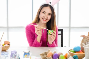 Paskalya bayramı konsepti, tavşan kulağı takan mutlu Asyalı genç kadın beyaz oda arka planında renkli Paskalya yumurtalarıyla Paskalya için yumurta boyuyor.
