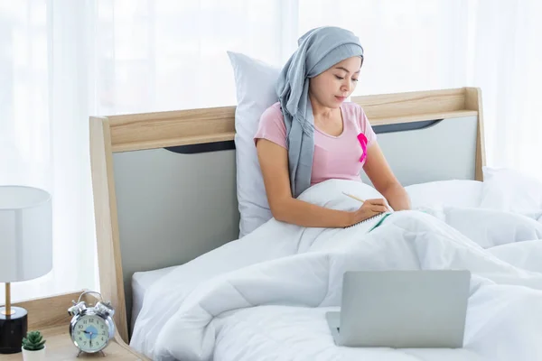 a asian women disease mammary cancer patient with pink ribbon wearing headscarf After treatment to chemotherapy with Take notes in a notebook and laptop on bed In the bedroom at the house,healthcare