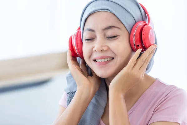 Happy a asian women disease mammary cancer patient with pink ribbon wearing headscarf in headphones is listening to music After treatment to chemotherapy sit on bed In the bedroom at the house