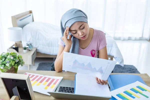 asian women disease mammary cancer patient in pink ribbon wearing headscarf After treatment to chemotherapy Have a headaches with working document business plan and laptop in office at home.