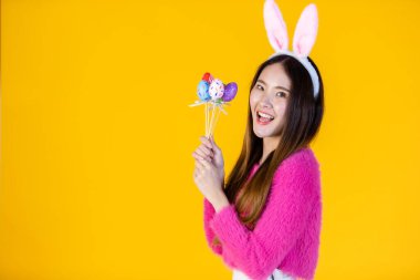 Paskalya tatili konsepti, mutlu gülümseyen Asyalı genç kadın tavşan kulaklı renkli Paskalya yumurtalarını tutan şiş sarı boş fotokopi stüdyosu arka planında izole edilmiş..