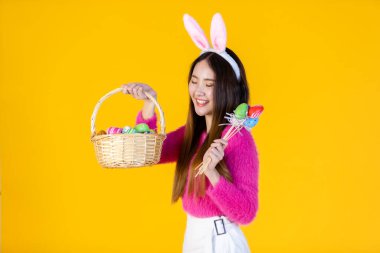 Paskalya bayramı konsepti, Mutlu Asyalı genç kadın tavşan kulaklı, elinde renkli Paskalya yumurtaları olan bir sepetle sarı boş fotokopi stüdyosu arka planında izole edilmiş..