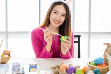 Paskalya bayramı konsepti, tavşan kulağı takan mutlu Asyalı genç kadın beyaz oda arka planında renkli Paskalya yumurtalarıyla Paskalya için yumurta boyuyor.