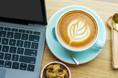 Kahve fincanında sütlü sütlü sütlü sütlü sütlü sıcak kahve ve iş konsepti sırasında kafedeki ahşap ofis masasında dizüstü bilgisayarlı ev yapımı muzlu kek.