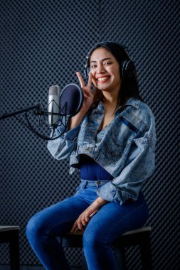 Profesyonel bir stüdyoda mikrofon önünde şarkı kaydeden, kulaklık takan genç Asyalı kadın vokalistin mutlu bir gülümsemesi.