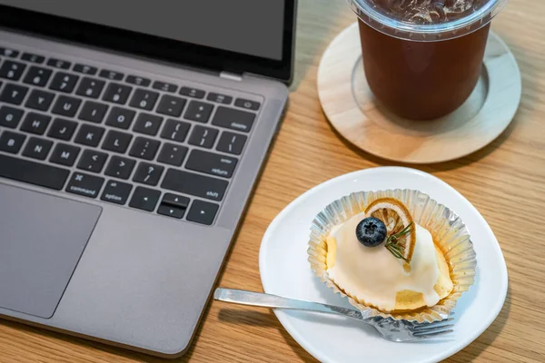 Close-up of ice coffee in cup mug and Homemade white orange cake with keyboard  laptop computer on wood desk office desk in coffee shop at the cafe,during business work
