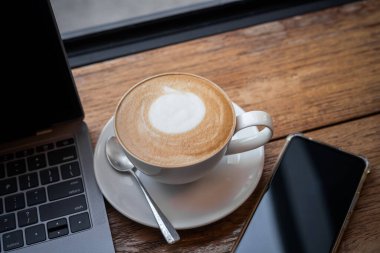 Kahve fincanında sütlü sütlü sütlü sütlü sütlü sütlü sütlü latte ve iş konsepti sırasında kafedeki ahşap ofis masasında akıllı telefon.