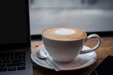 Kahve fincanında sütlü sütlü sütlü sütlü sütlü sütlü sütlü latte ve iş konsepti sırasında kafedeki ahşap ofis masasında akıllı telefon.