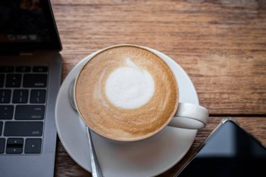 Kahve fincanında sütlü sütlü sütlü sütlü sütlü sütlü sütlü latte ve iş konsepti sırasında kafedeki ahşap ofis masasında akıllı telefon.