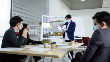 Asyalı bayan antrenör koruyucu maske hoparlörü, ofisteki COVID-19 odasında çeşitli cinsiyetlerle (LGBT) buluşan iş adamlarına ters yüz grafiği sunumu yapıyor 