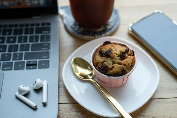 Close-up of Homemade Banana cup cake and ice coffee in cup mug with wireless earphones on laptop computer on wood desk office desk in coffee shop at the cafe,during business work concept
