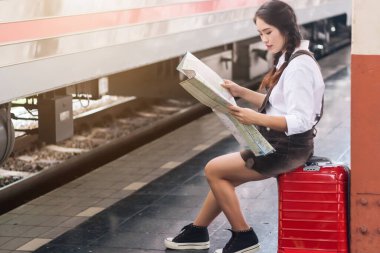 Bekleyen Asyalı hamile turist, bankta oturmuş tren istasyonunda kırmızı bavulla haritaya bakıyor..