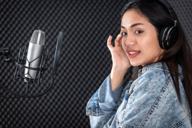 Profesyonel bir stüdyoda mikrofon önünde şarkı kaydeden, kulaklık takan genç Asyalı kadın vokalistin mutlu bir gülümsemesi.