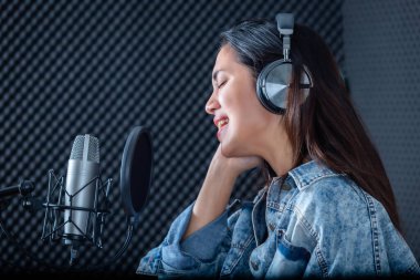 Profesyonel bir stüdyoda mikrofon önünde şarkı kaydeden, kulaklık takan genç Asyalı kadın vokalistin mutlu bir gülümsemesi.