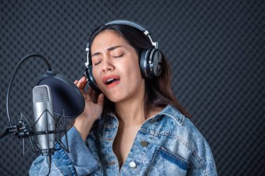 Profesyonel bir stüdyoda mikrofon önünde şarkı kaydeden, kulaklık takan genç Asyalı kadın vokalistin mutlu bir gülümsemesi.